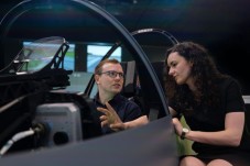 Man wearing glasses sitting inside a flight simulator while speaking with a colleague standing outside.