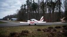 Small red and white aeroplane on an airfield runway surrounded by trees.