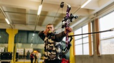 A man aiming a purple compound bow at a target in an indoor archery range.